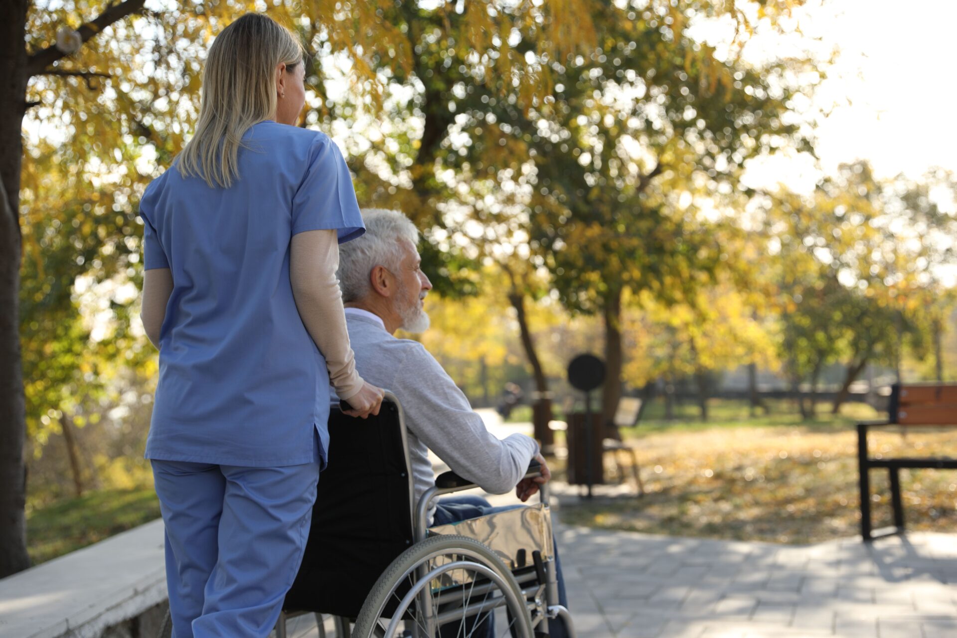 Caregiver assisting senior man on wheelchair in park, back view and space for text. Home health care service